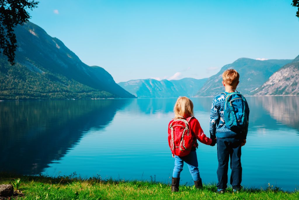 Jente med rød ryggsekk og gutt med blå ryggsekk ser ut mot fjord og fjell